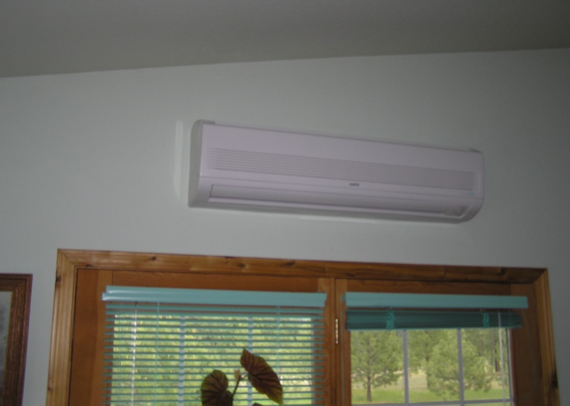 An indoor ductless heat pump unit mounted high on a wall above a wooden-framed window with blinds partially closed.